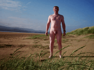 Nude man standing on a beach with grass in the foreground and hills in the background.