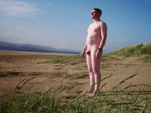 Nude man standing on a beach with grassy dunes and blue sky in the background.