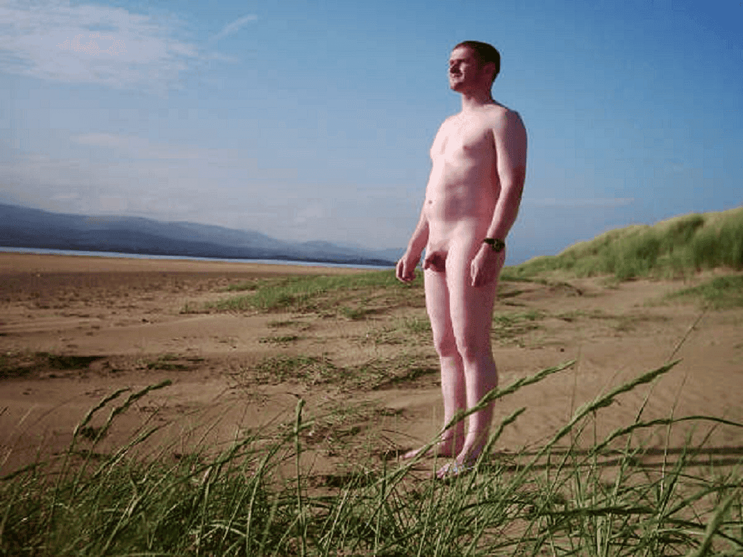 Nude man standing on a beach with grassy dunes and blue sky in the background.