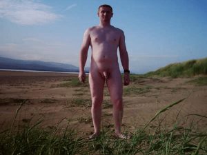 A man stands confidently on a sandy beach, surrounded by green grass and rolling hills. The background features a clear blue sky, creating a serene atmosphere. His stance reflects a relaxed demeanor, enjoying the tranquility of the natural setting.