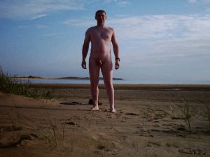 A man standing nude on a sandy beach, with a serene sea and sky in the background. He is positioned confidently, surrounded by gentle grasses swaying in the breeze, as soft clouds float above in a bright blue sky.