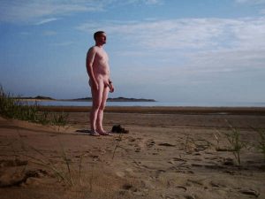 A nude man stands alone on a sandy beach, gazing at the tranquil sea under a vast, blue sky dotted with soft clouds. The golden sand stretches around him, with beach grasses swaying gently in the light breeze, creating a serene, idyllic atmosphere.