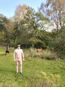 Naked man standing outdoors in a garden with trees and lush greenery, wearing a hat and sneakers.