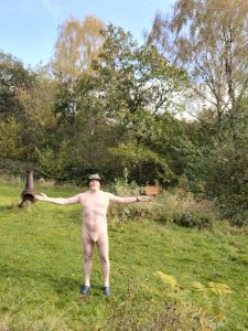 Outdoor nude portrait of a man standing with arms outstretched in a garden setting.