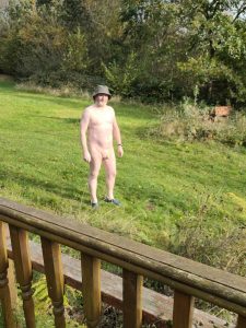 Nude man standing in a garden with trees and greenery in the background.