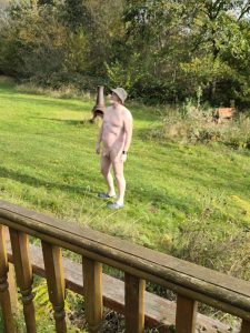Naked man standing in a lush garden with trees and greenery in the background.