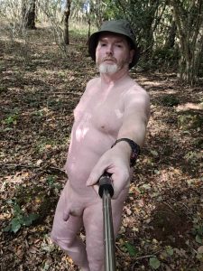 Nude man taking a selfie in a wooded outdoor setting.