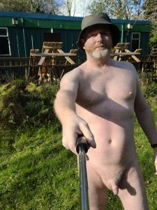 Nude man taking a selfie outdoors in a garden setting.