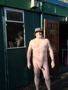 Naked man standing outdoors in front of a green building, wearing a hat and watch, with a reflection.