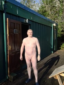 Naked man standing outside a green cabin in natural sunlight.