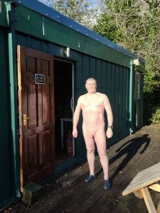 Naked man standing outside a green shipping container on a wooden deck.