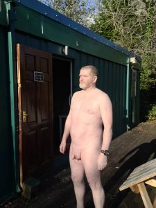 Naked man standing outside a green cabin, enjoying the outdoors in natural sunlight.