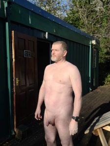 Nude man standing outdoors near a green building with trees in the background.