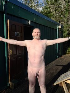 Naked man standing outside with arms wide open in front of a green building.
