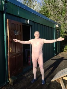 Naked man standing outside a green container with arms outstretched.