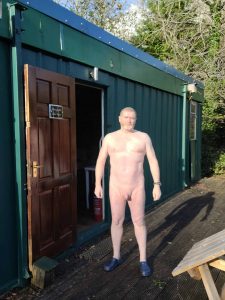Naked man standing outside a green cabin in a natural outdoor setting.