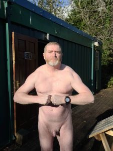 Naked man standing outside a green shed, flexing his muscles in a confident pose.