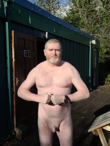 Man standing nude outside a green building with trees in the background.