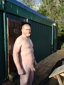 Nude man standing outside a green building with trees in the background.