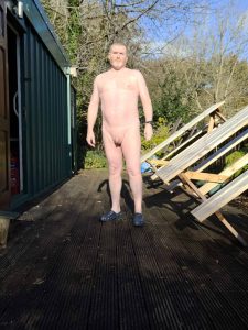 Naked man standing outside on a wooden deck with trees in the background.
