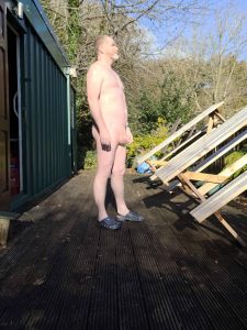 Naked man standing on a wooden deck outdoors with trees in the background.