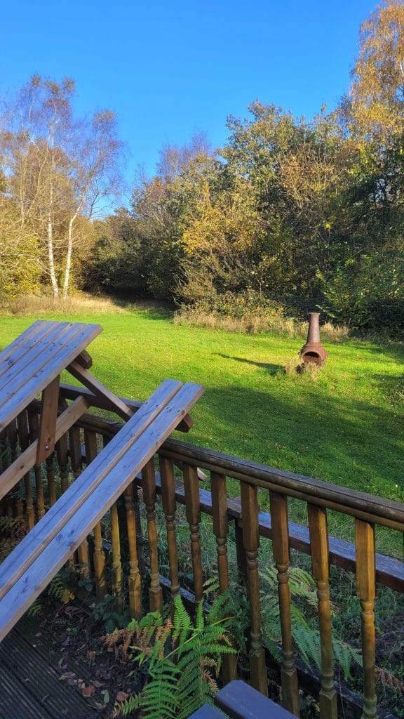 A wooden deck with several angled planks overlooks a green lawn surrounded by trees. In the background, there is a metal chiminea and clear blue sky.