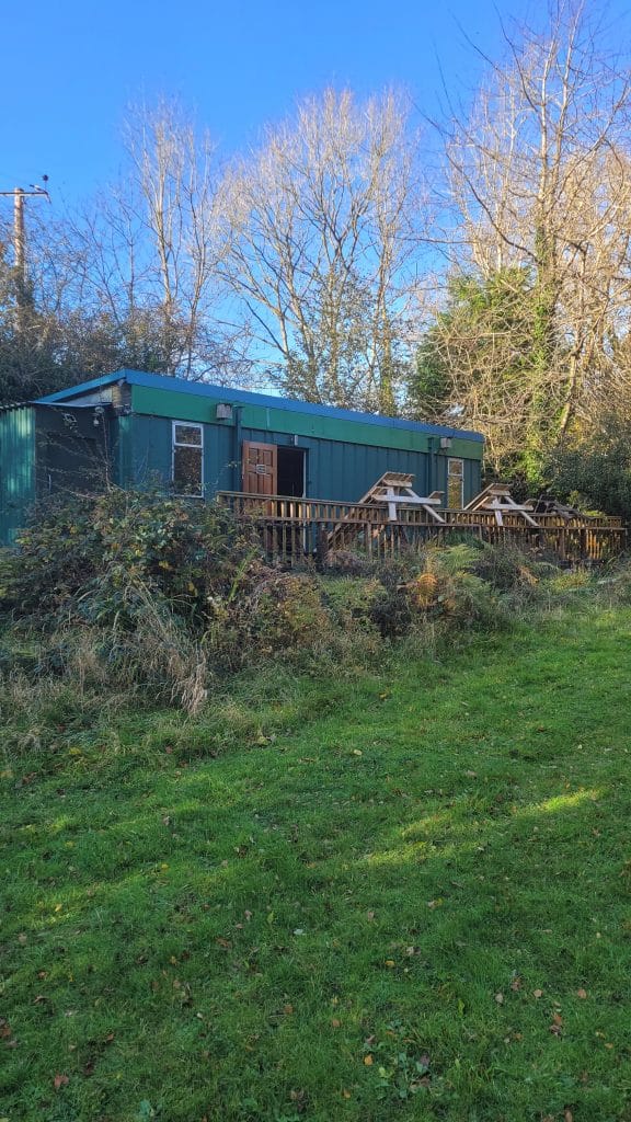 A green cabin surrounded by trees and shrubs, with a wooden deck in front, located in a grassy area under a clear blue sky.