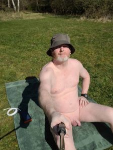 A middle-aged man sitting on a green towel in a sunlit grassy area, wearing a grey bucket hat and sunglasses. He is mostly nude, with a relaxed posture, holding a selfie stick to capture the moment. Surrounding him is a tranquil landscape of trees and open fields under a clear blue sky.