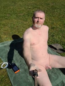 A man sitting on a green towel on grass, holding a selfie stick. He has short grey hair and a beard, and is naked, enjoying the sunlight. A solar charger and a hat can be seen nearby, indicating a relaxed day outdoors.
