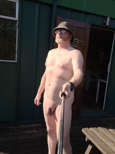 Nude man with hat and sunglasses taking a selfie outside.