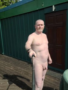 Nude man standing outside, taking a selfie with a selfie stick.