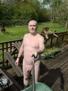 Naked man taking a selfie outdoors on a wooden deck with greenery in the background.