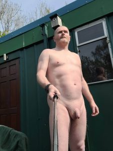 Man standing outdoors, completely nude, holding a selfie stick in front of a green building.