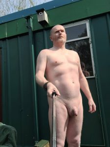 Naked man standing outdoors with a walking stick in front of a green building.