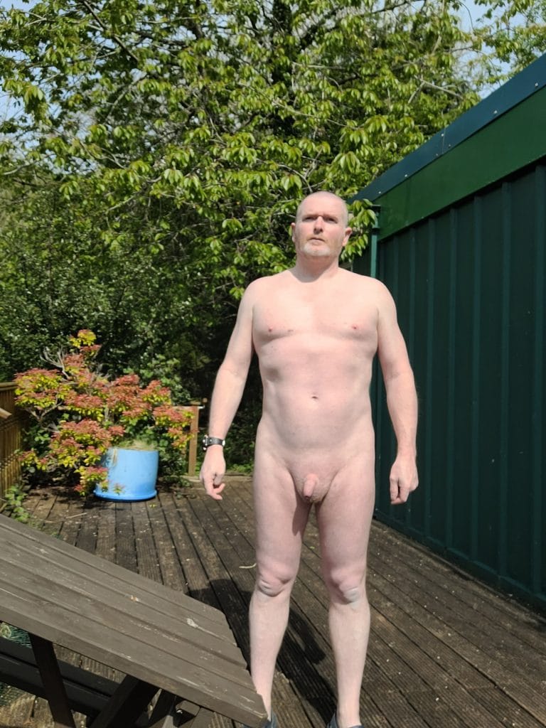 A person standing outdoors in a natural setting, surrounded by greenery and wooden decking. The individual is facing the camera, and there are various plants and a blue container in the background.