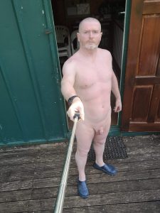 Nude man taking a selfie outside a pub, standing on wooden decking.