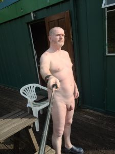 Naked man standing outside with selfie stick on wooden deck.