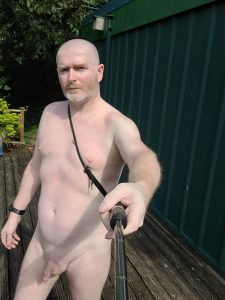 Nude man taking a selfie outdoors with a selfie stick in a garden setting.