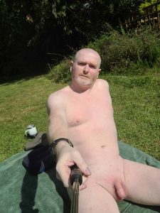 Naked man relaxing outdoors on a sunny day, taking a selfie with a selfie stick.