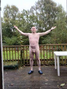 Nude man standing outdoors with arms raised on a wooden deck, surrounded by trees.