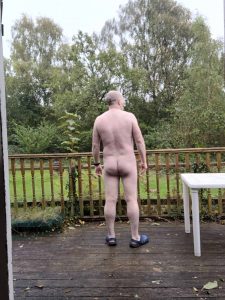 Naked man standing on a wooden deck, facing away, with a garden and trees in the background.