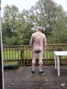 Naked man standing on a wooden deck overlooking a garden with trees.
