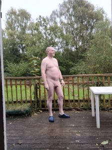 Naked man standing on a wooden deck with a garden and trees in the background.