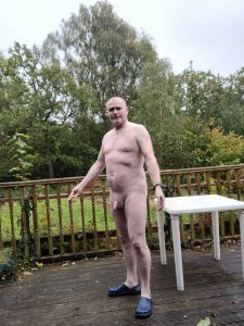 Naked man standing on a wooden deck with trees in the background.