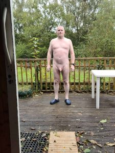 Naked man standing on a wooden deck outdoors, surrounded by greenery and trees.