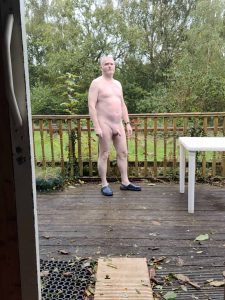 Naked man standing outdoors on a wooden deck with greenery in the background.