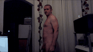 Man standing shirtless in a room with curtains and electronic equipment.