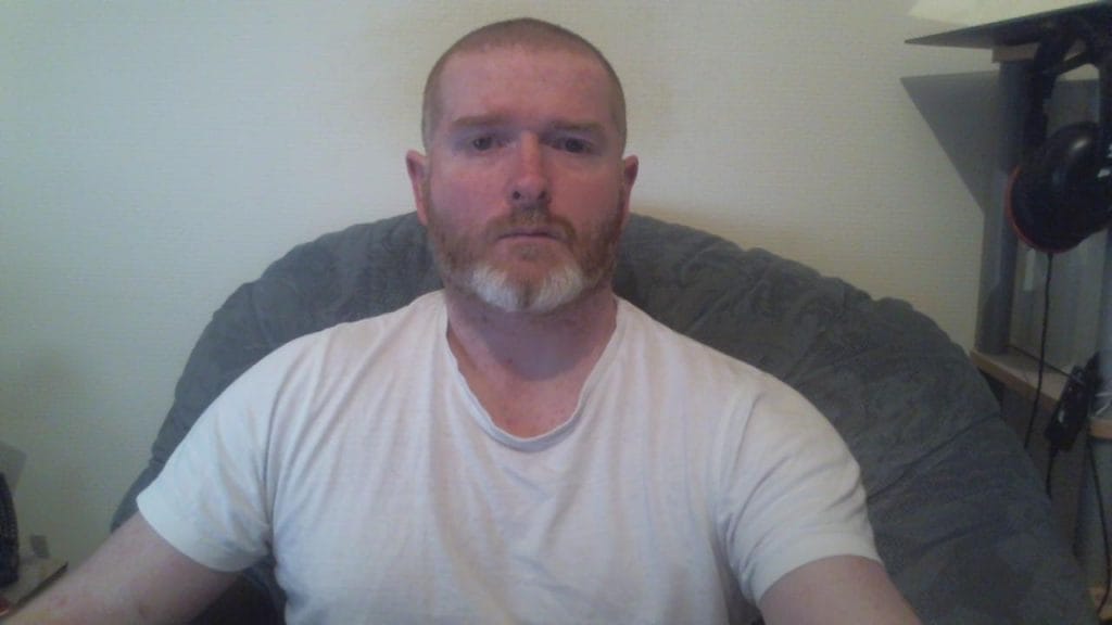 A man with a short beard and shaved head, wearing a plain white T-shirt, sits in a large, plush chair. The background shows a simple room with a few objects, creating a casual atmosphere.